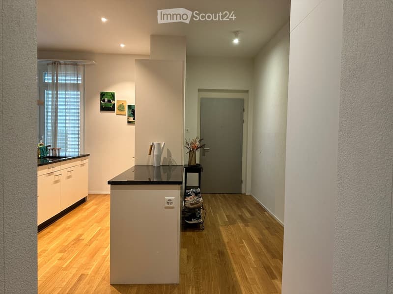 A modern hallway with wooden flooring, white walls, and a small kitchen area visible on the left. The hallway features a closed door at the end and a small table with shoes and a vase of flowers near the door.