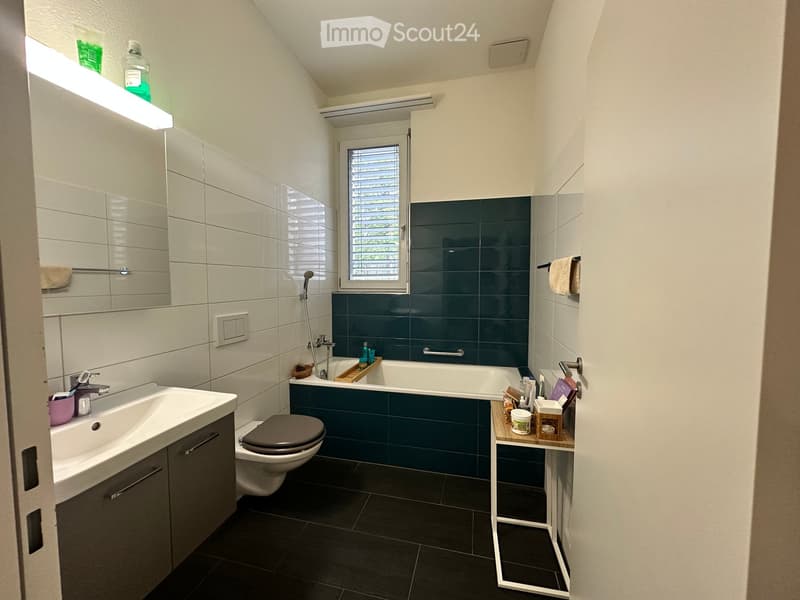 Modern bathroom with white tiles, dark blue accent wall, bathtub, toilet, sink, and mirror. Features a window with blinds and a small shelf with toiletries.