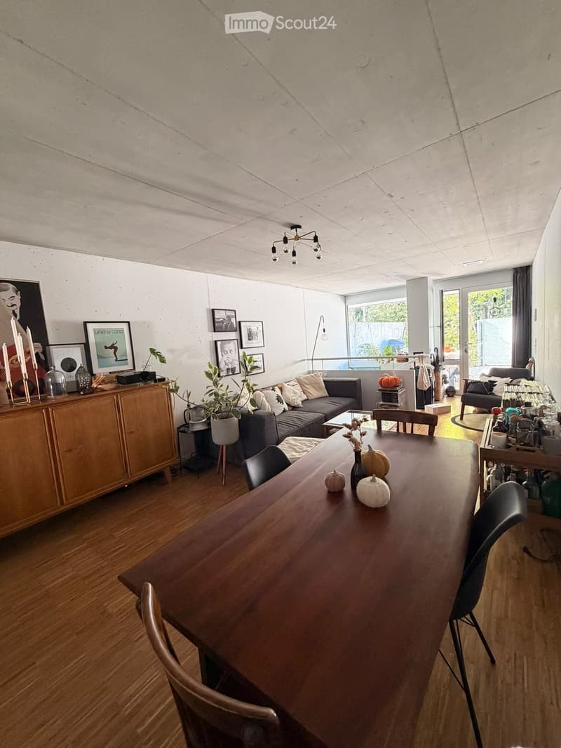 Modern living and dining area with wooden flooring, exposed concrete ceiling, large windows, and a view of the outdoor space. Features include a grey sectional sofa, wooden dining table with chairs, a wooden cabinet with decorative items, and several pott