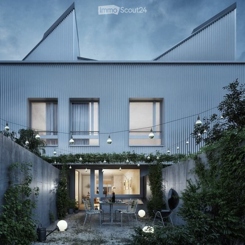 Modern two-story house with a sleek, grey exterior and large windows. The outdoor area features a small patio with a dining table, chairs, and string lights, surrounded by greenery and plants.