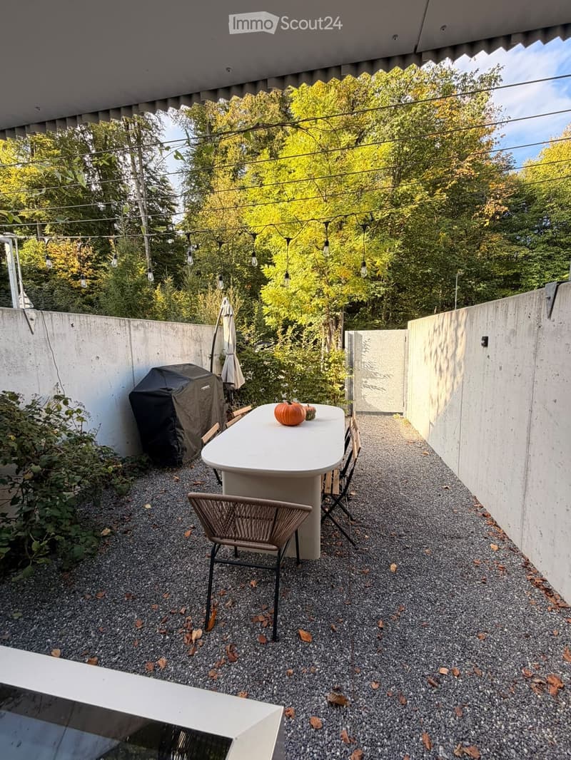 Compact outdoor patio with a white table, chairs, a covered grill, and string lights, surrounded by concrete walls and greenery.