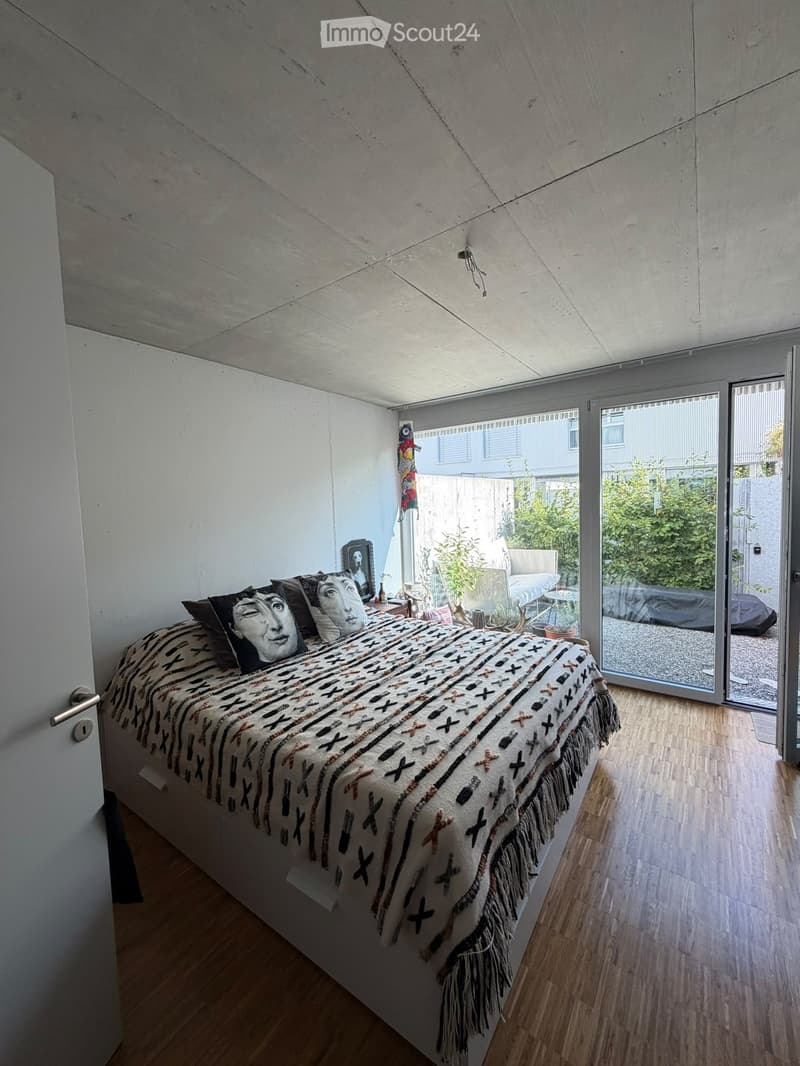 Modern bedroom with wooden flooring, large bed with patterned bedspread and pillows, nightstand, mirror, and a sliding glass door leading to a patio area.