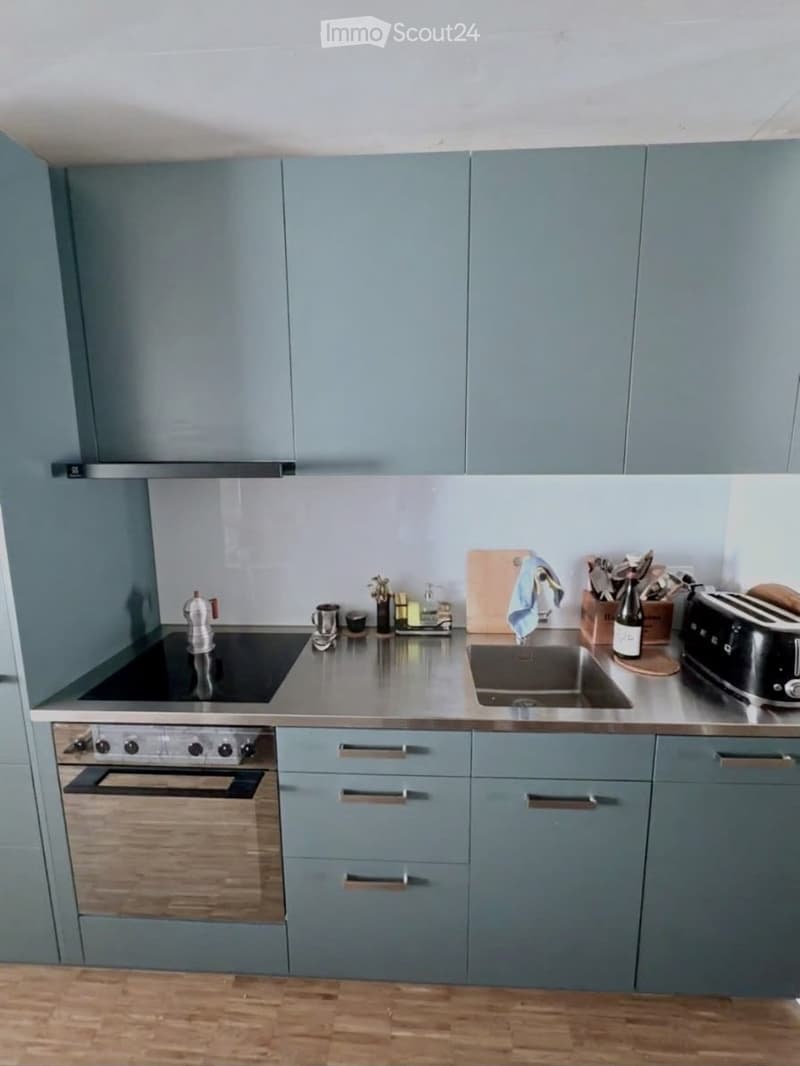 Modern kitchen with grey cabinets, stainless steel countertops, and a wooden floor. Features a stove, oven, sink, toaster, and various kitchen utensils.