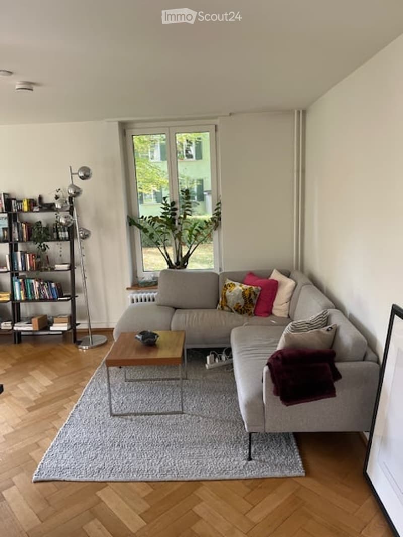 Modern living room with a grey sectional sofa, wooden coffee table, bookshelf, floor lamp, and a large plant near the window.