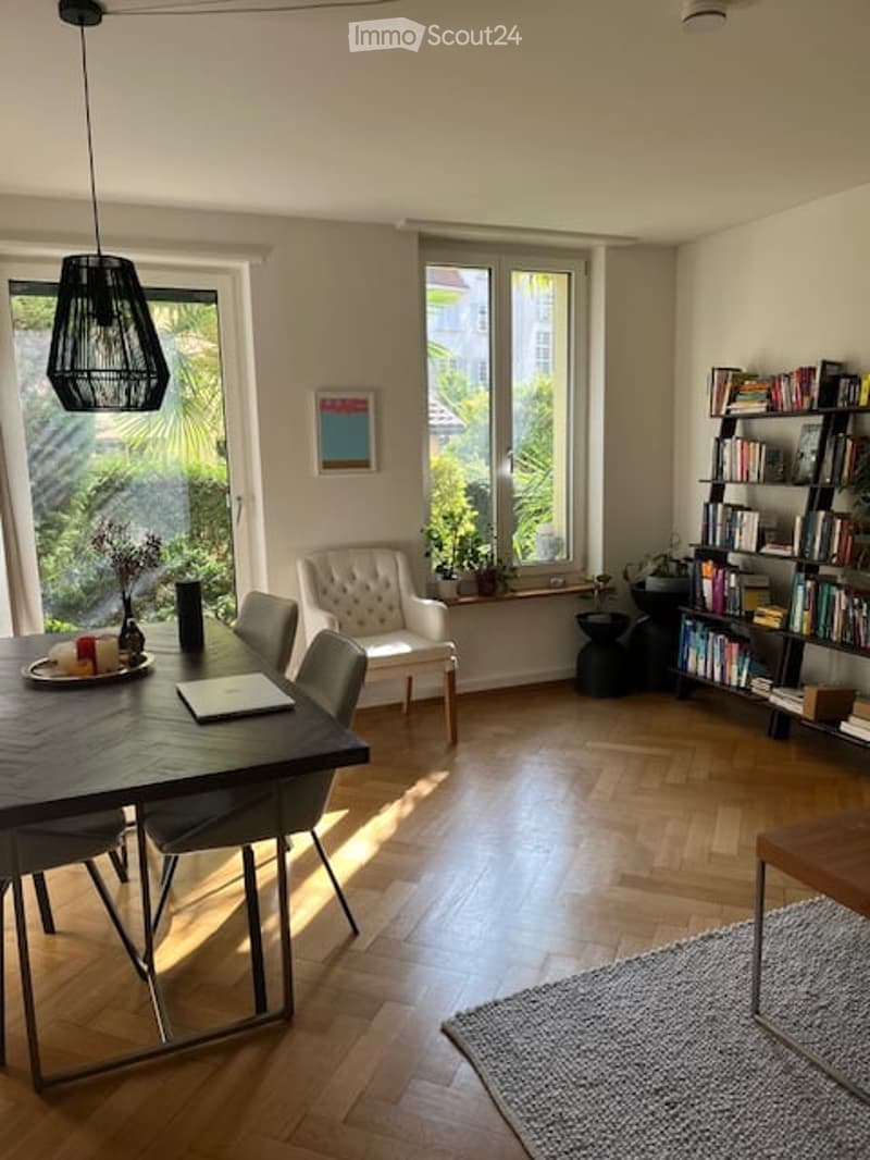 Bright, modern dining area with wooden flooring, large windows, a dark wooden dining table, chairs, a white armchair, a bookshelf, and a floor lamp.
