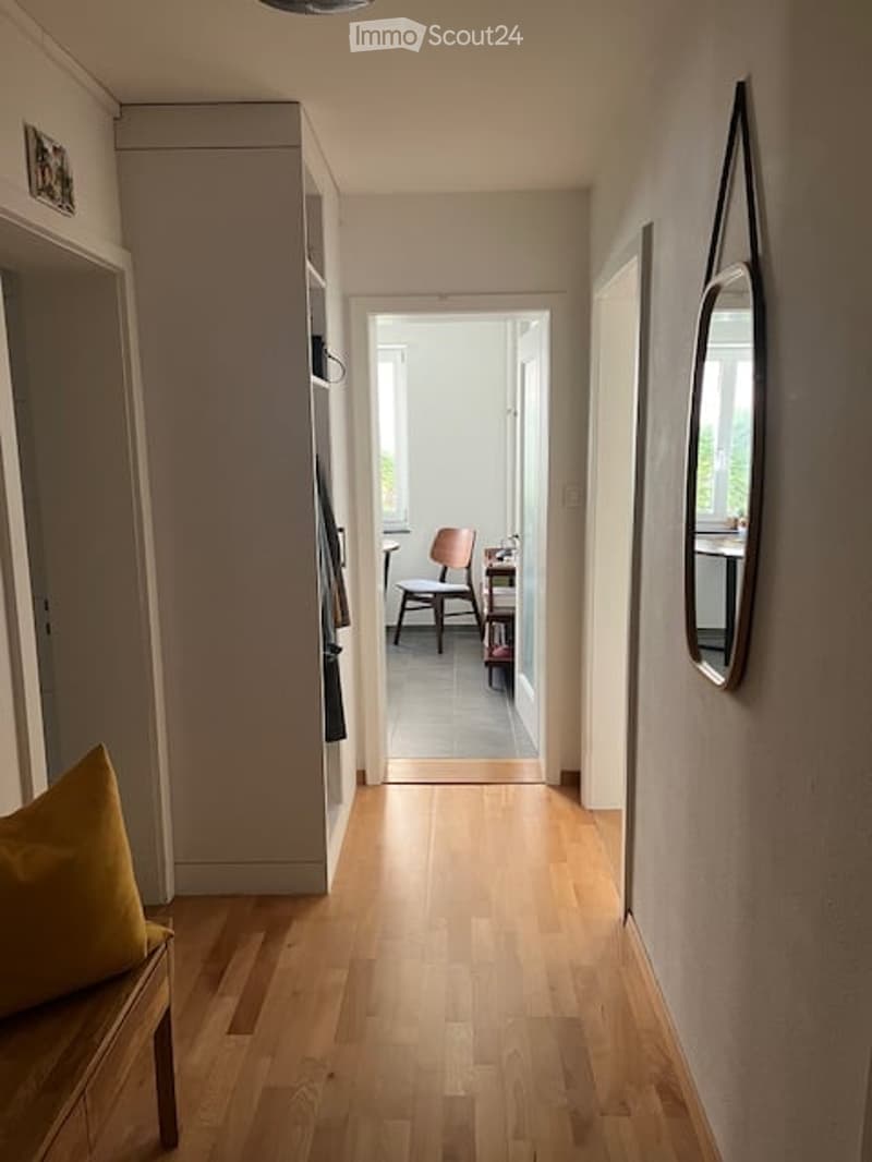 A narrow hallway with light wooden flooring, white walls, and a bench with a yellow cushion on the left side. A mirror is mounted on the right wall, and at the end of the hallway, a small room is visible with a chair and a table.