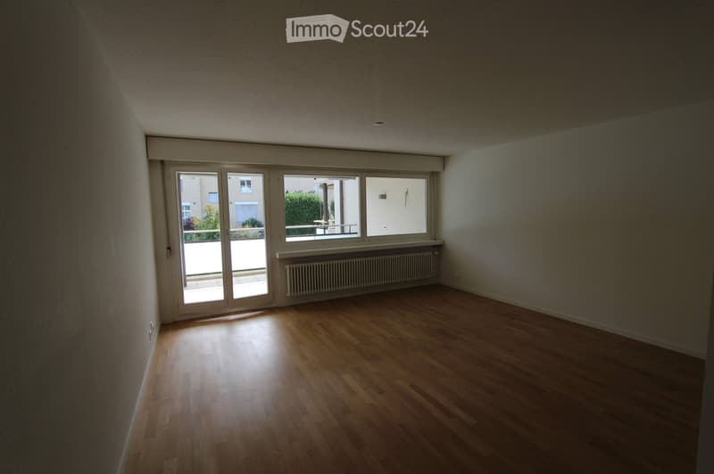 Empty room with wooden flooring, white walls, large windows, and a radiator under one window.