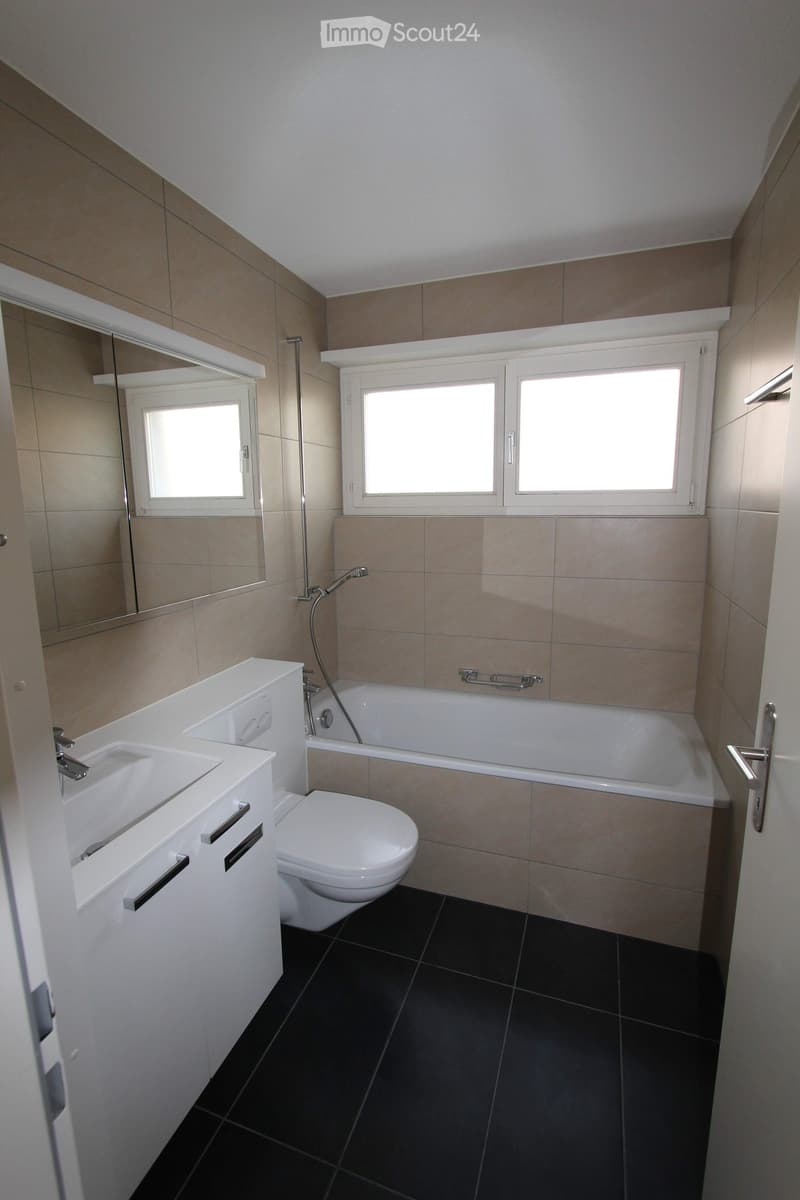 Modern bathroom with a white sink, toilet, bathtub, and large mirror. Features dark tiled flooring and beige tiled walls.