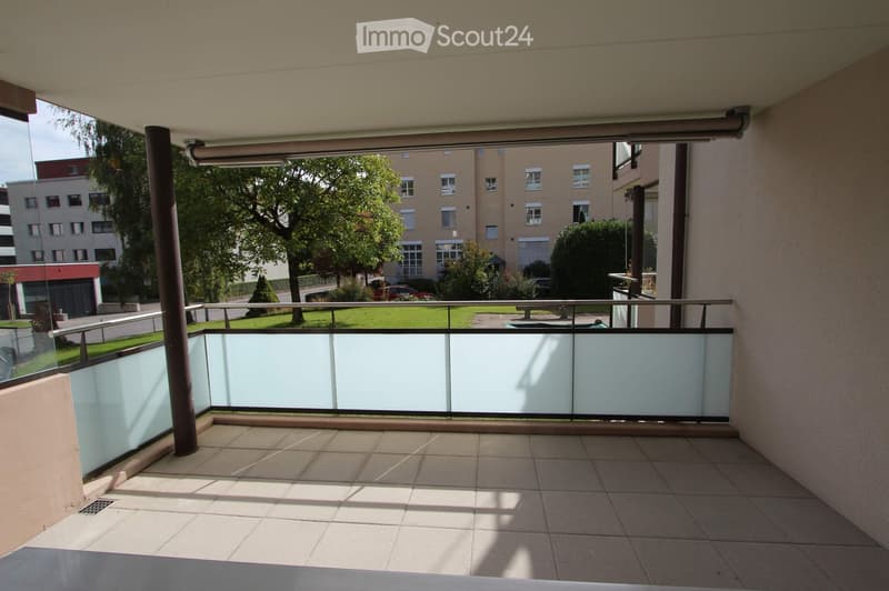 Balcony with tiled floor, glass railing, and view of a green area with trees and buildings.