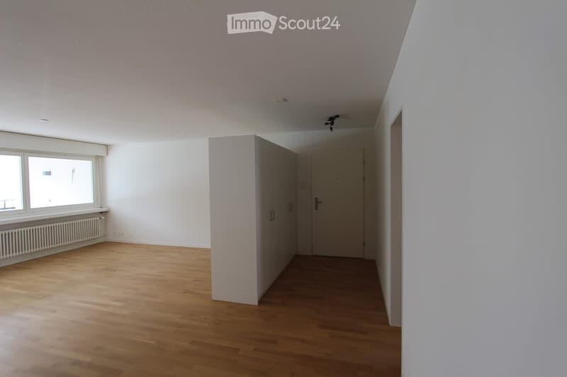 Empty room with wooden flooring, white walls, a window with a radiator below, and a door leading to another space.