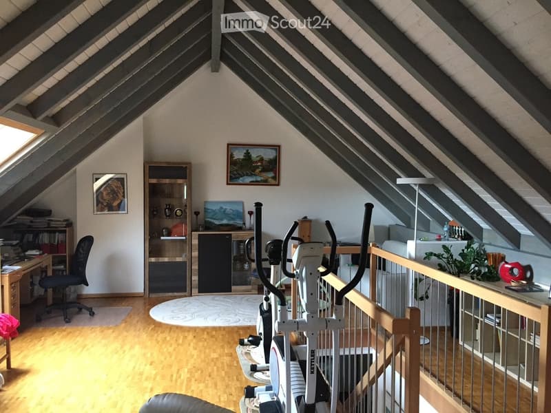 Spacious attic room with wooden flooring, slanted ceilings, and large windows. Features a desk with a chair, bookshelf, exercise bike, and a small seating area with a stool and painting on the wall.