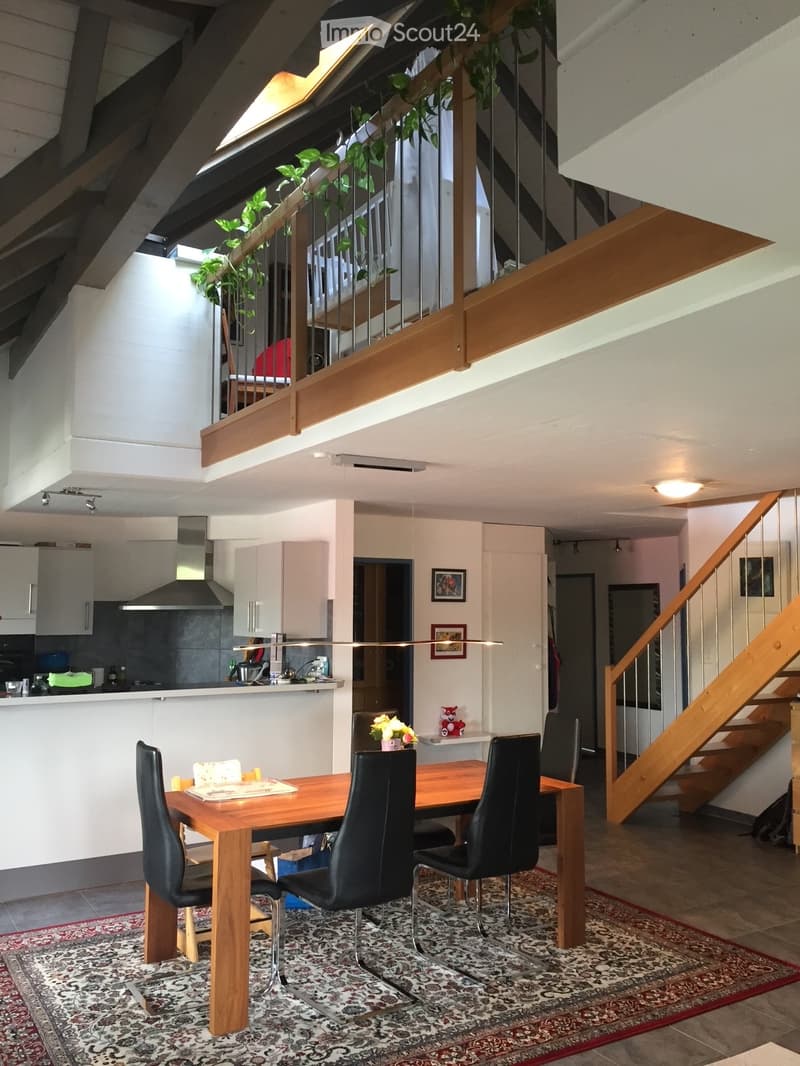 Open-plan living and dining area with a wooden dining table, black chairs, modern kitchen, staircase leading to an upper level, and decorative elements like a flower vase and picture frames.