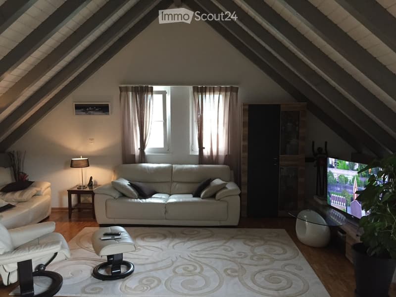 Bright living room with slanted wooden ceiling, large window with curtains, white sofa, armchair, ottoman, coffee table, lamp, TV, and potted plant.