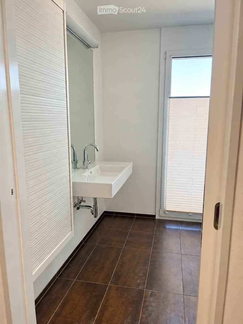 Modern bathroom with a white sink, chrome faucet, and dark brown tiled floor. Features a large mirror and a window with blinds.