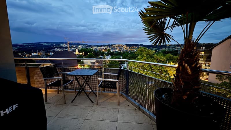 Balcony with a cityscape view, featuring a small table, chairs, and a potted palm tree.