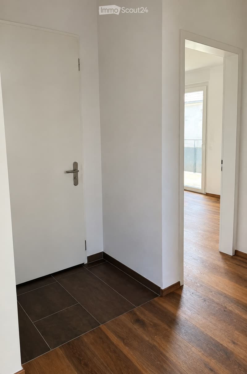 Empty hallway with white walls, dark tiled floor, and wooden flooring leading to an open area with a view of a balcony.