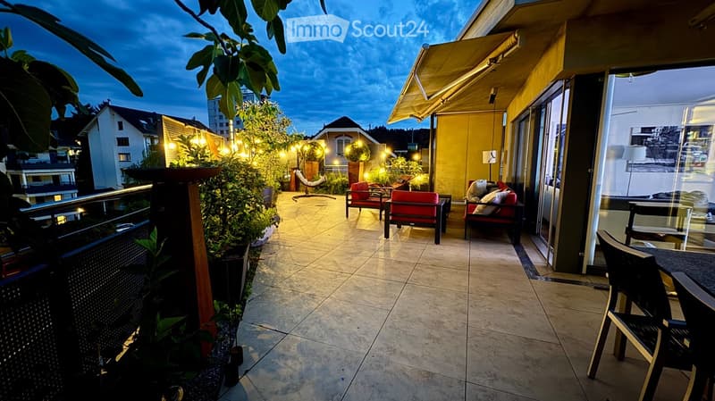 Balcony with tiled floor, outdoor furniture including chairs, a small table, and a sofa, surrounded by potted plants and string lights.