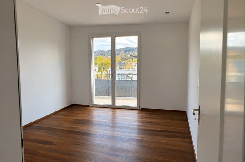 Empty room with wooden flooring, white walls, and a large sliding glass door leading to a balcony with a view of trees and a building.