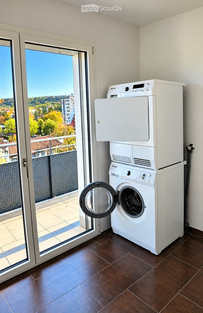 Modern laundry room with dark wooden flooring, white washing machine and dryer stacked, large window with view of cityscape and greenery.