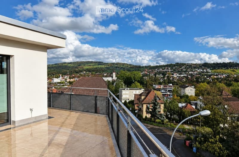 Balcony with tiled floor, metal railing, and a view of a town with houses, trees, and hills under a partly cloudy sky.