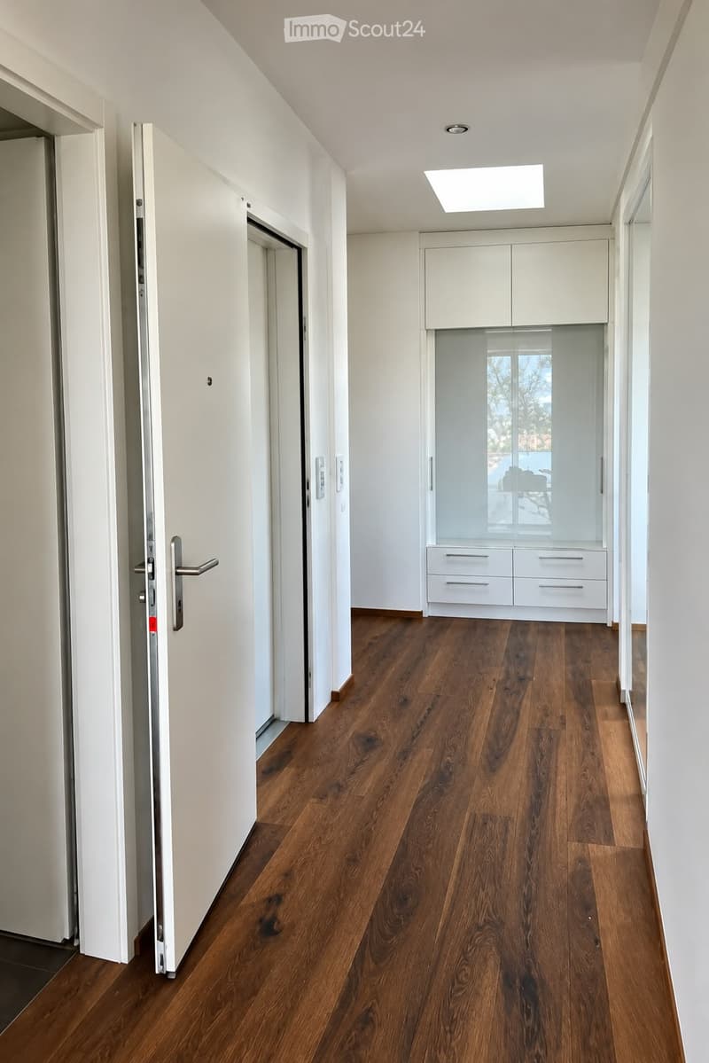 Modern hallway with wooden flooring, white walls, and built-in storage units with glass doors. Features recessed lighting and a partially open door leading to another room.
