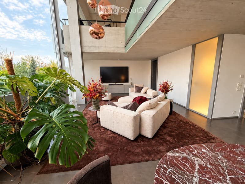 Modern living room with large windows, white sofa, coffee table, ottoman, armchair, marble side table, television, and decorative plants.
