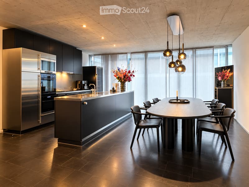 Modern kitchen with stainless steel appliances, dark cabinets, a central island, and a round dining table with chairs. Features pendant lighting and large windows with sheer curtains.