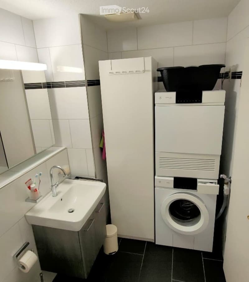 Bathroom with a sink, mirror, refrigerator, and a stacked washing machine and dryer unit.