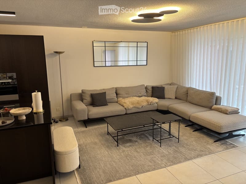 Modern living room with a grey sectional sofa, coffee table, ottoman, and a floor lamp. A large mirror hangs on the wall, and a small table holds decorative items.