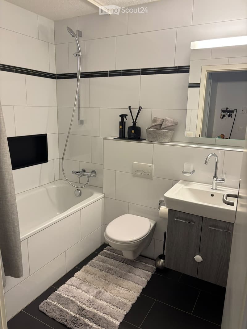 Modern bathroom with white tiles, bathtub, toilet, sink, mirror, and a grey towel on the floor.