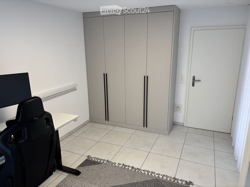 Modern, minimalist room with a white tiled floor, a grey carpet, a white table, a black chair, and a large screen mounted on the wall. The room features built-in grey cabinets and a white door.