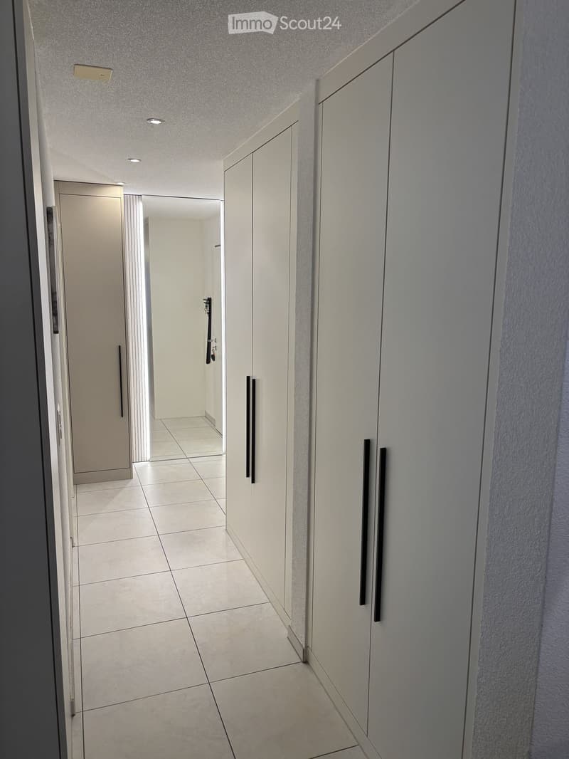 Clean, modern hallway with white tiled flooring, white walls, built-in cabinets with black handles, and recessed ceiling lighting.