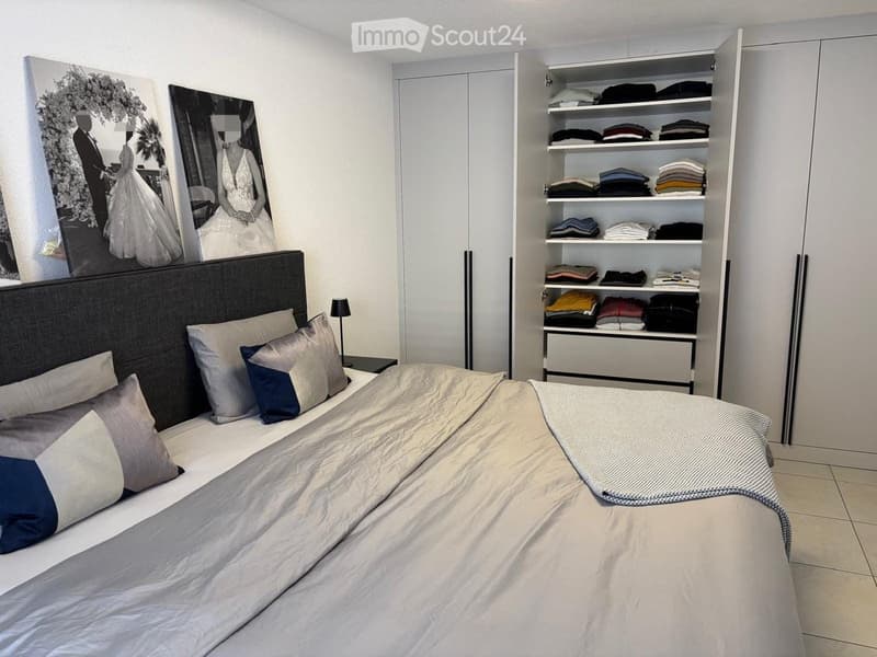 Modern bedroom with a large bed, decorative pillows, a blanket, a wardrobe with neatly folded clothes, a nightstand with a lamp, and two large picture frames above the bed.
