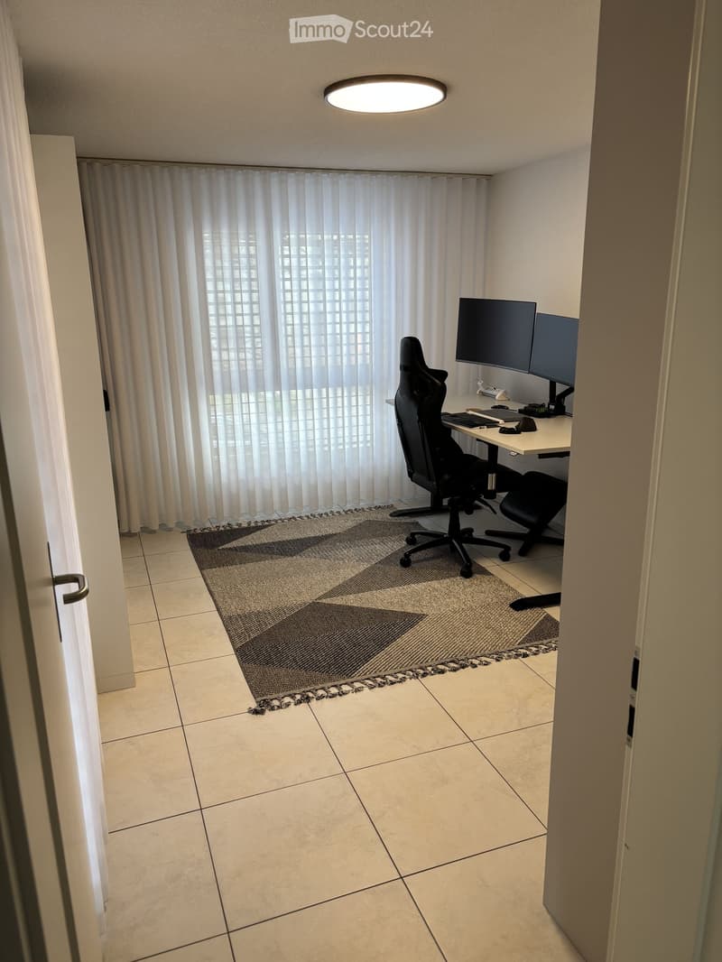 A modern, furnished room with a desk setup, two monitors, a chair, and a large window with curtains.