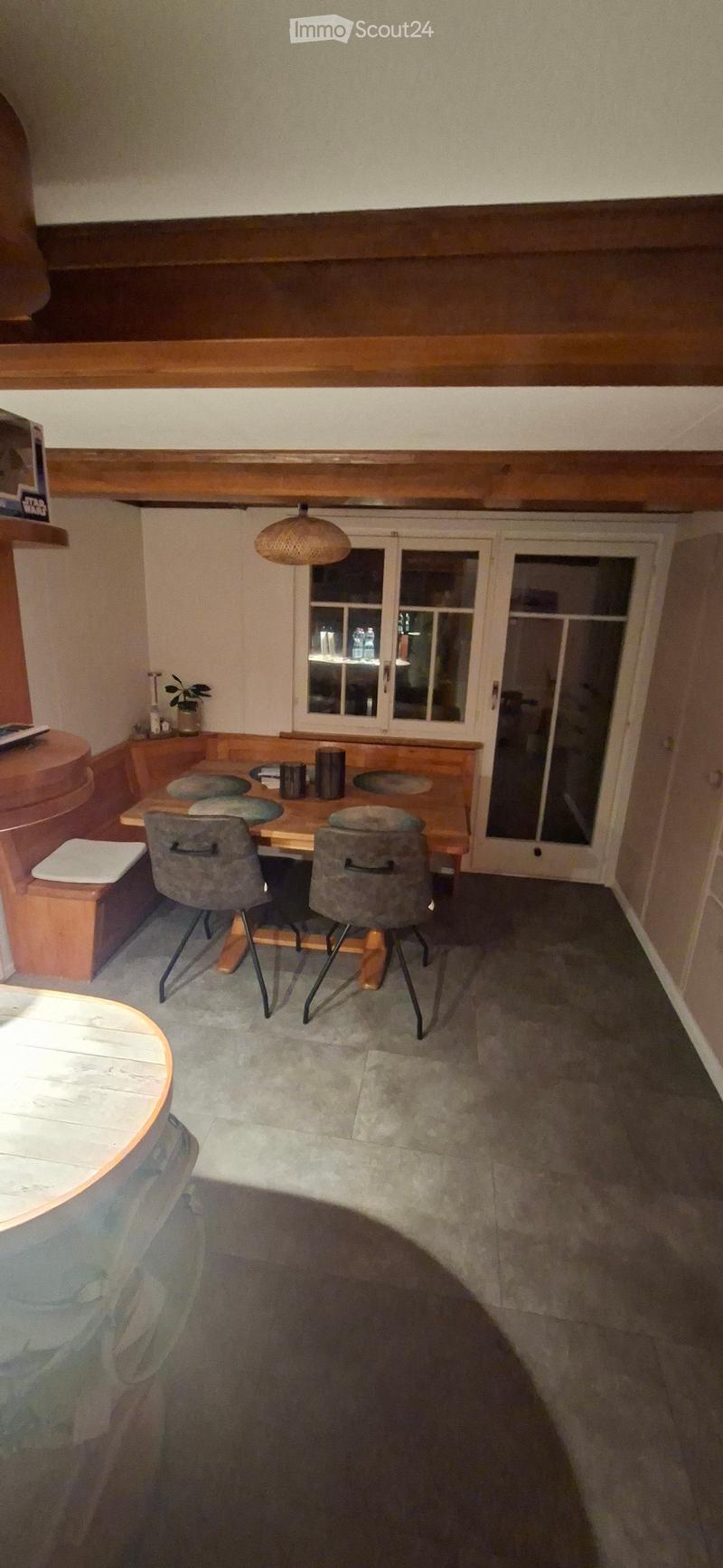 Dining area with a wooden table, two chairs, a bench, and a small round table with a stool nearby. The room features exposed wooden beams on the ceiling and a large window with glass doors leading outside.