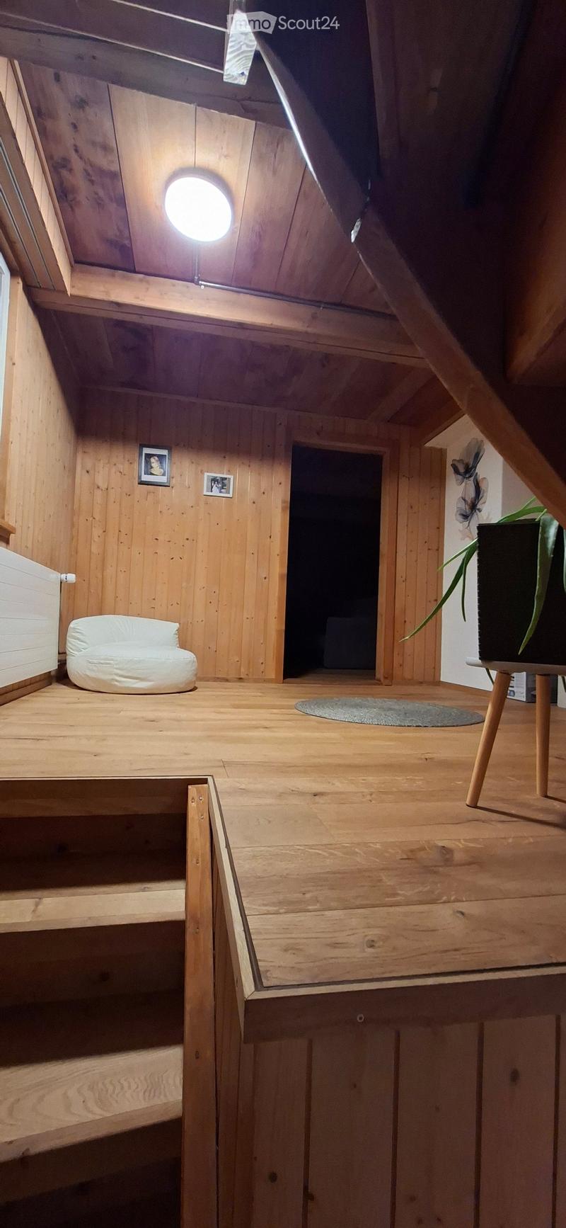 A cozy, wooden-furnished attic room with a small white sofa, a round wooden table with a plant, and a few picture frames on the wall. The room features exposed wooden beams and a skylight.