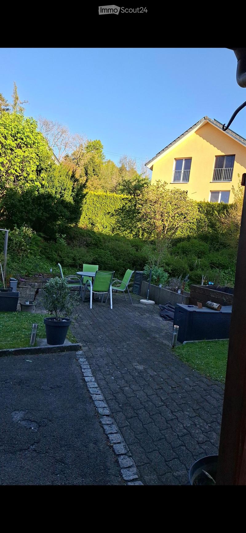 Garden area with a paved pathway, small seating area with a table and chairs, potted plants, and a yellow house in the background.