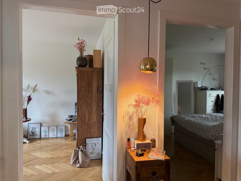 A modern hallway leading to a bedroom with a bed, nightstand, and decorative items. The hallway features a wooden floor, a hanging lamp, and a small table with a vase and books.