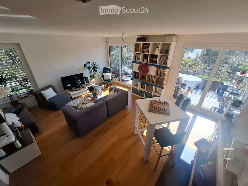 Bright living room with wooden flooring, large windows, and a view of the outdoors. Features a grey sectional sofa, coffee table, bookshelf, and a small dining area with a table and chairs.