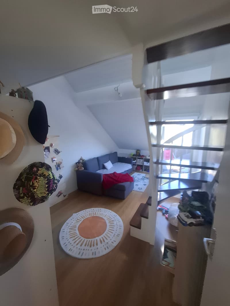 A cozy living room with a grey sofa, a small table, a round white stool, and a staircase leading to another level. The room features wooden flooring and white walls with some decorations.
