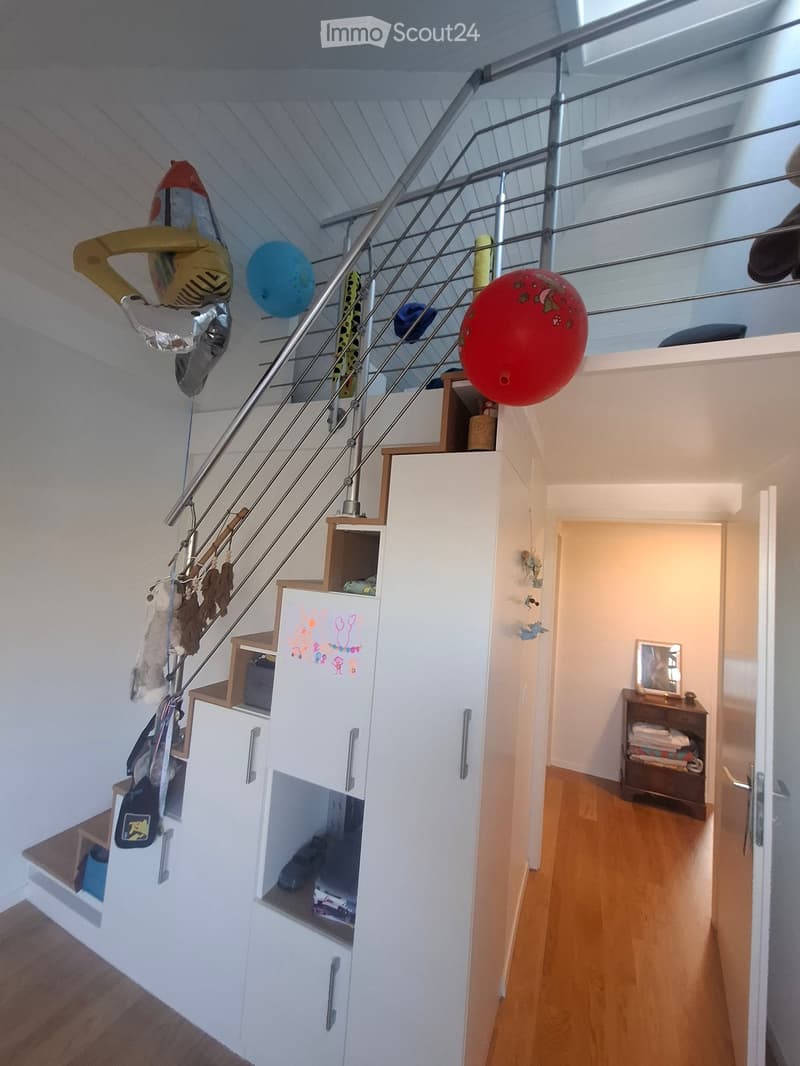 Modern hallway with wooden flooring, white walls, staircase with metal railing, and various children's toys and decorations hanging on the railing. A small cabinet and a shelf with folded towels are visible in the adjacent room.