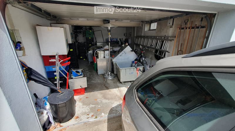 Garage with a car parked inside, various storage items including boxes, tools, and a bicycle, with a heater lamp in the center.
