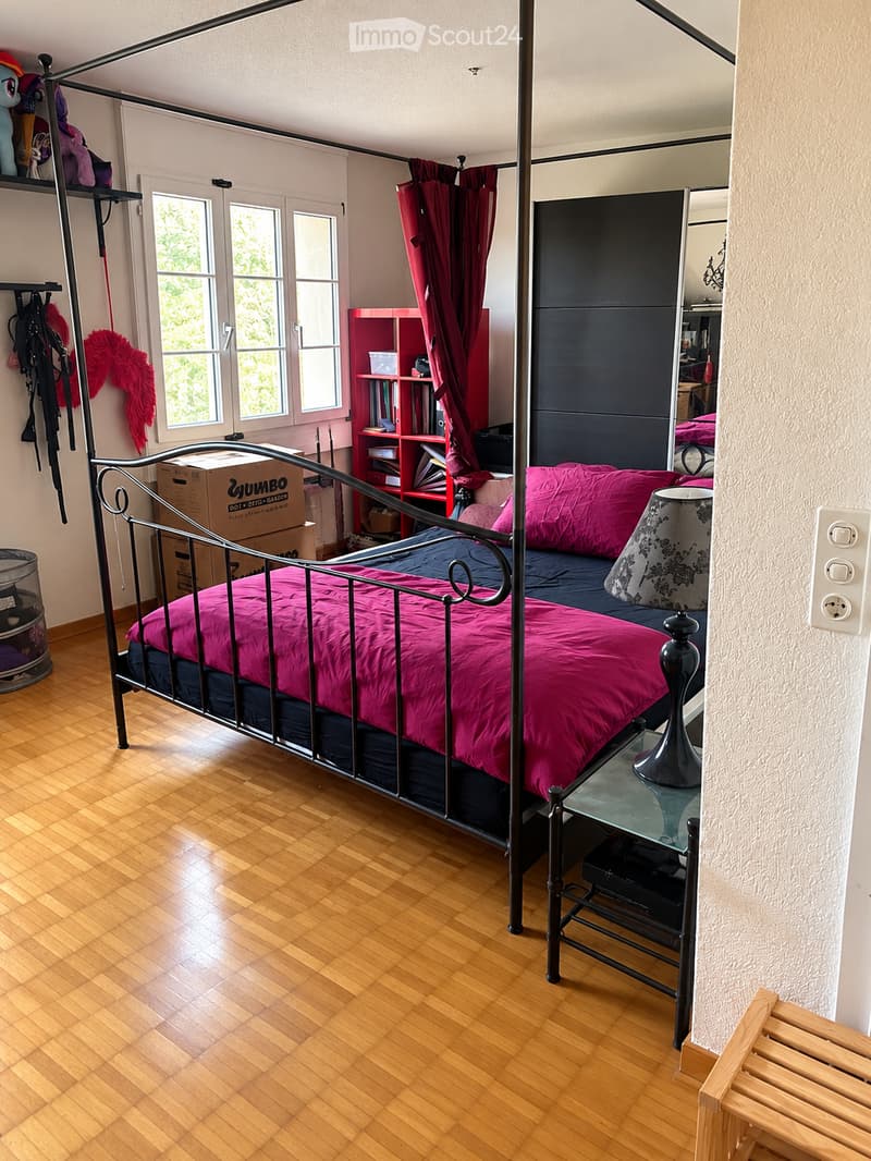 A cozy bedroom with wooden flooring, a metal-framed bed with pink bedding, a nightstand with a lamp, a red bookshelf, and a wooden bench near the door.