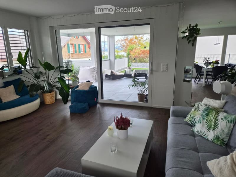 Modern living room with wooden flooring, large sliding glass doors leading to a balcony, grey sofa with green and white cushions, white coffee table with a small plant, blue round chair, and additional seating area on the balcony.