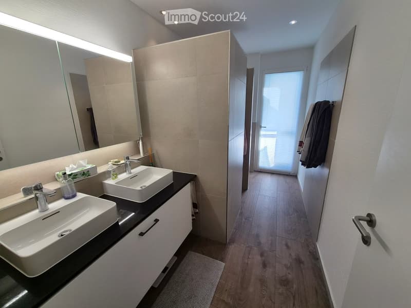 Modern bathroom with two white sinks, sleek faucets, large mirror, and a towel hanging on the wall. The space features light-colored tiles and a wooden floor.