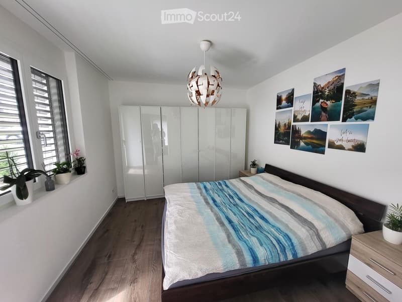 Modern bedroom with a large bed, a wardrobe, nightstand, potted plants, and picture frames on the wall.