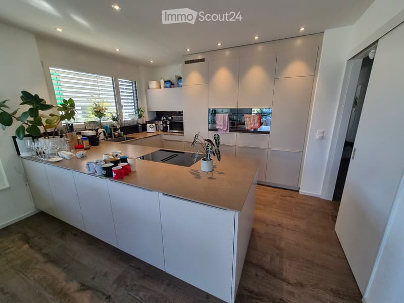 Modern kitchen with white cabinets, a central island with a wooden countertop, and various appliances including a sink, oven, microwave, toaster, and coffee maker. The island is topped with cups, bowls, wine glasses, and a small plant. The kitchen feature