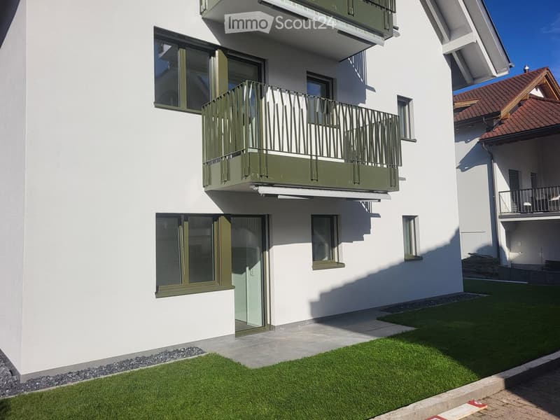 Modern two-story building with white exterior, green-framed windows, and a balcony on the upper floor. The ground floor has a large window and a door leading to a small garden area with artificial grass.