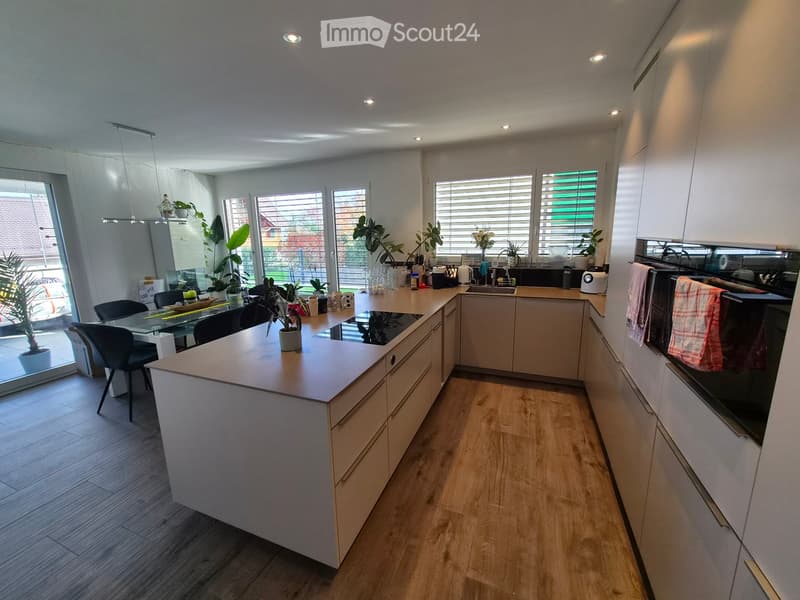 Modern kitchen with light wooden cabinets, a central island, and a dining area with a glass table and chairs. The space features a range of appliances, including an oven, microwave, and fridge, along with various kitchen items and potted plants.