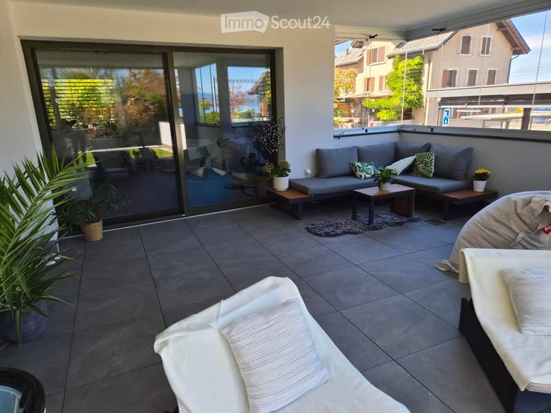 Modern balcony with grey sofa, small wooden table, white lounge chairs, and potted plants, offering a scenic view of the surroundings.